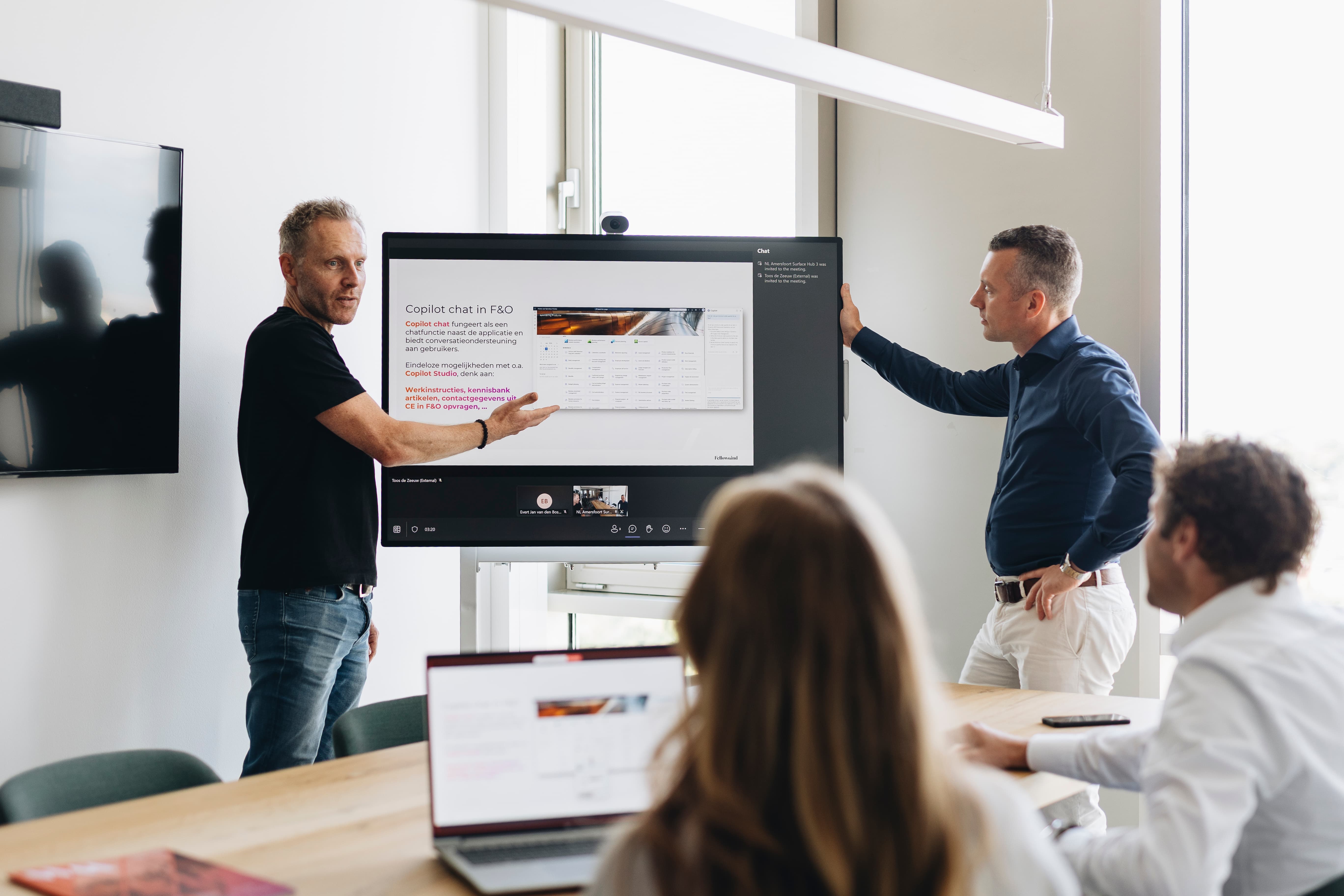Zakelijke presentatie met twee mannen bij een groot scherm in een moderne vergaderruimte, met laptop en deelnemers aan tafel