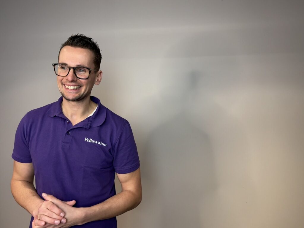 Man met paarse polo en bril staat tegen een effen grijze achtergrond