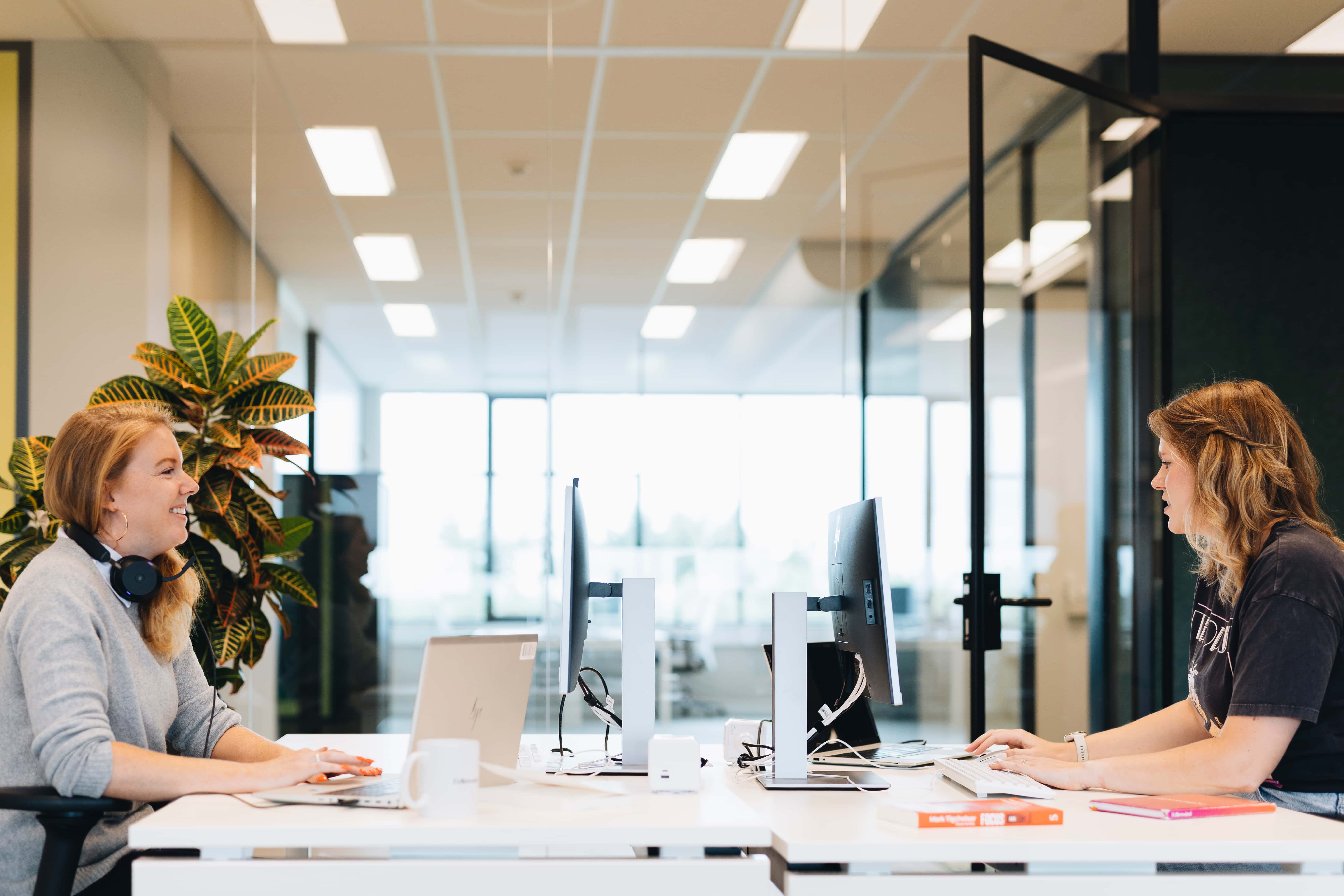 Twee vrouwen werken tegenover elkaar aan een bureau in een moderne kantoorruimte met computers en glazen wanden