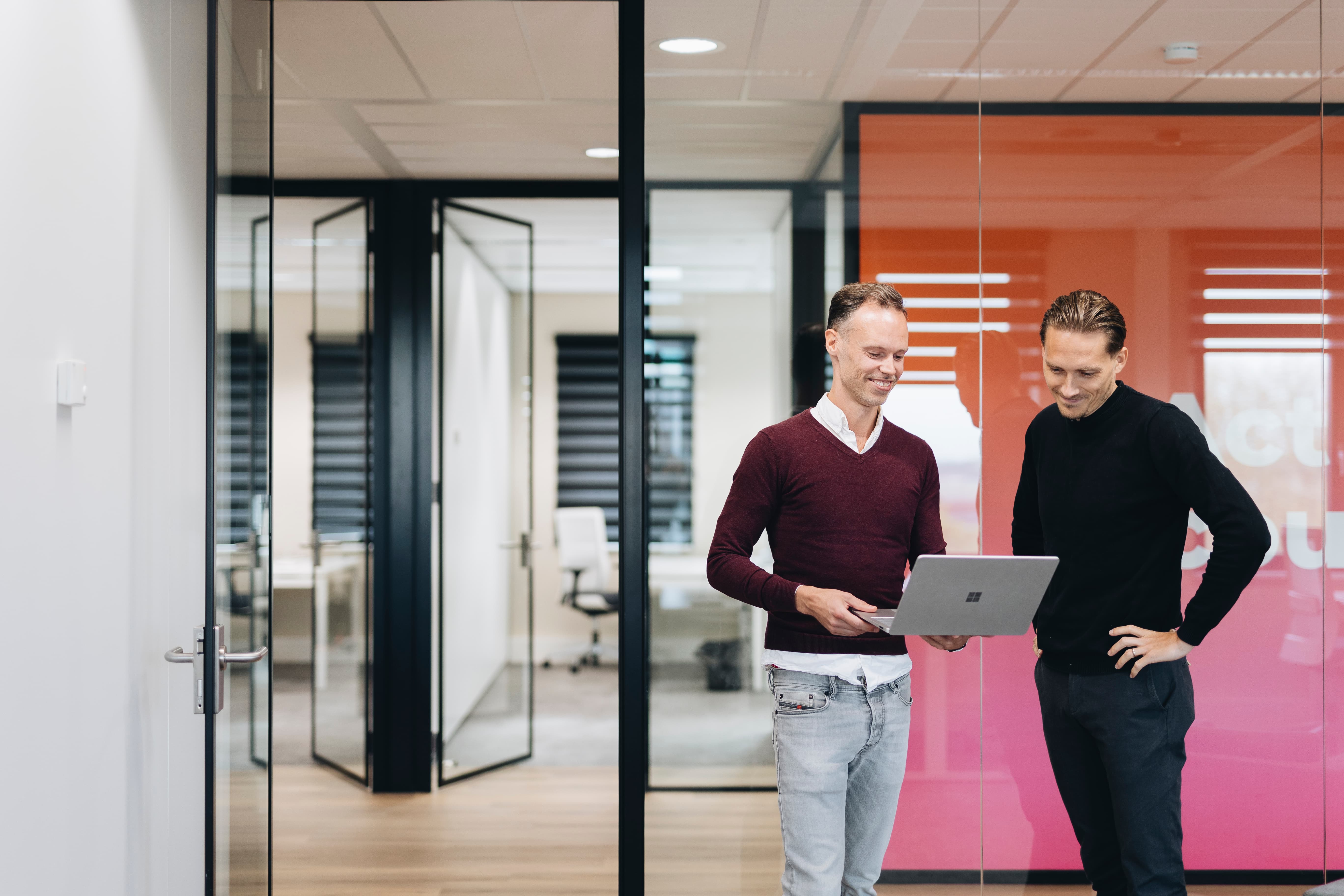 Twee collega’s overleggen in een moderne kantoorruimte met laptop en glazen vergaderruimtes
