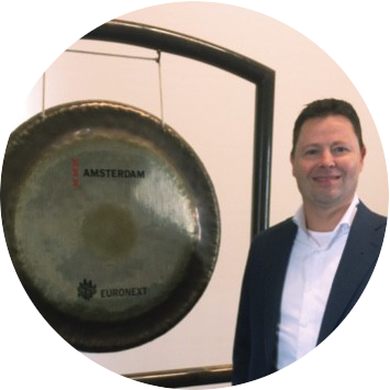 Een man in een donker pak staat naast een grote gong met Amsterdam- en Euronext-opdruk, tijdens een zakelijk evenement
