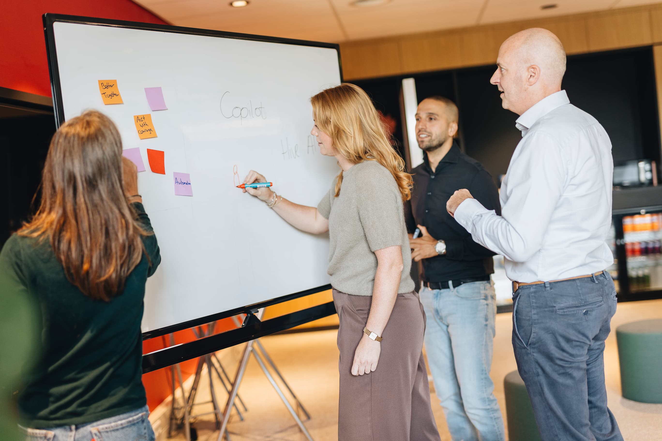 Teamoverleg met vier personen bij een whiteboard, waarbij ideeën en notities worden besproken en opgeschreven