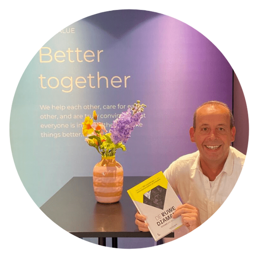 Man met wit overhemd houdt een boek vast naast een tafel met bloemen, voor een paarsblauwe muur met de tekst Better together