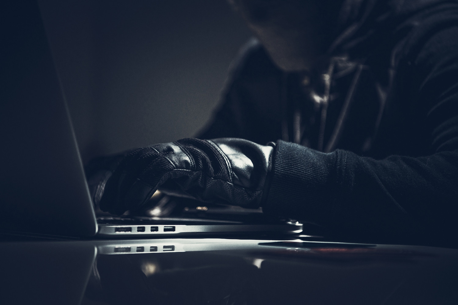 Persoon in donkere kleding en handschoenen typt op een laptop in een schemerige setting, met focus op cyberbeveiliging en online privacy