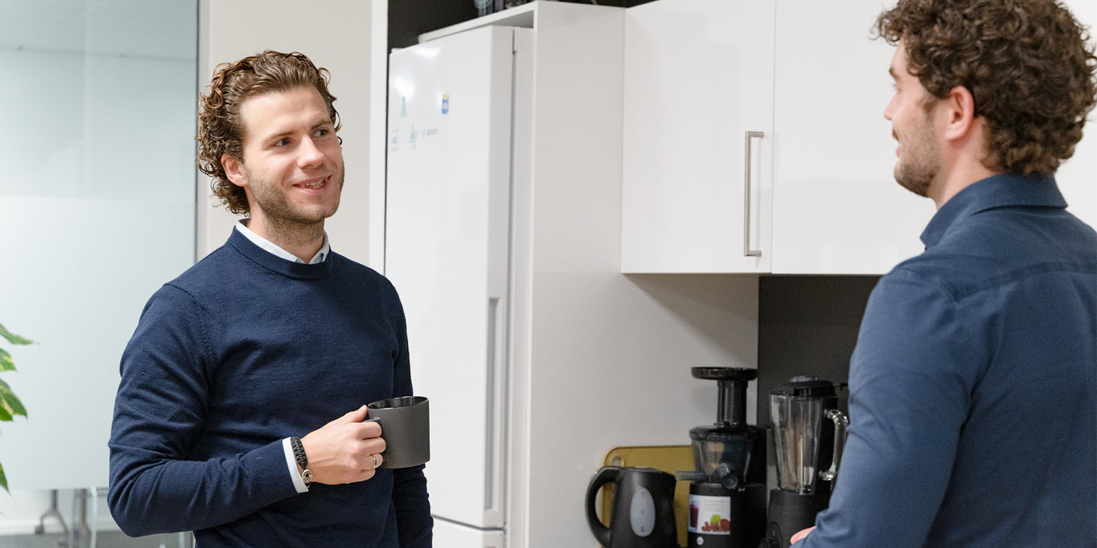 To mænd taler med hinanden i et lille kontor-køkken. Den ene har en Fellowmind-kaffekop i hånden.