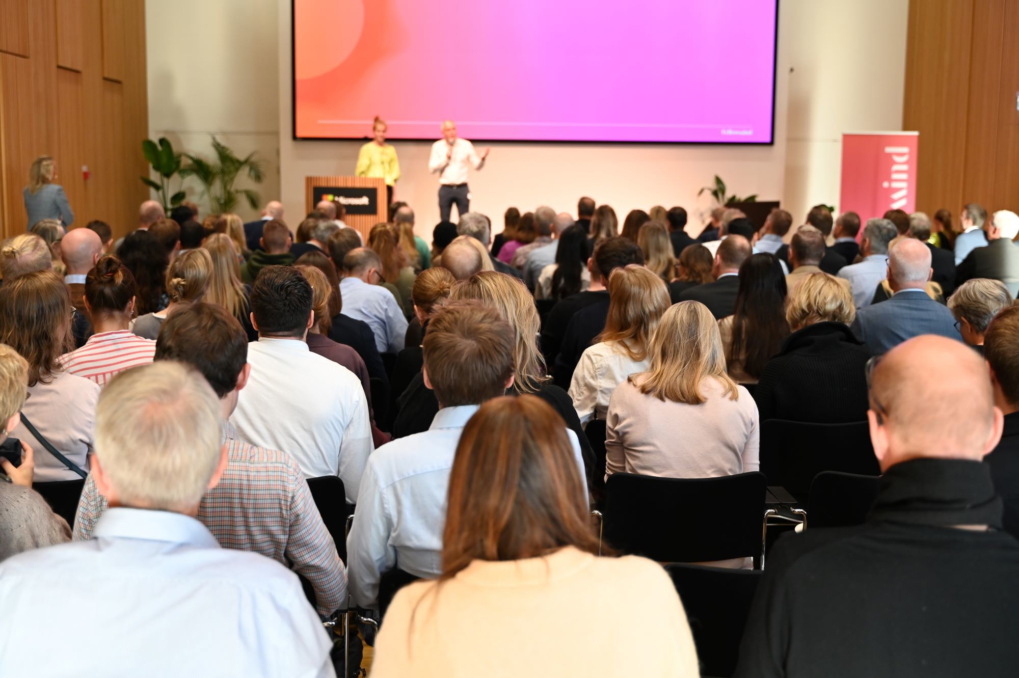 Publik på föreläsning i konferenssal med två talare på scen och stor presentationsskärm i bakgrunden