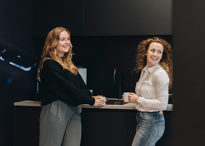 Twee vrouwen staan in een moderne keuken en praten met een kopje koffie in de hand. Donkere keukenkasten en een minimalistisch interieur op de achtergrond