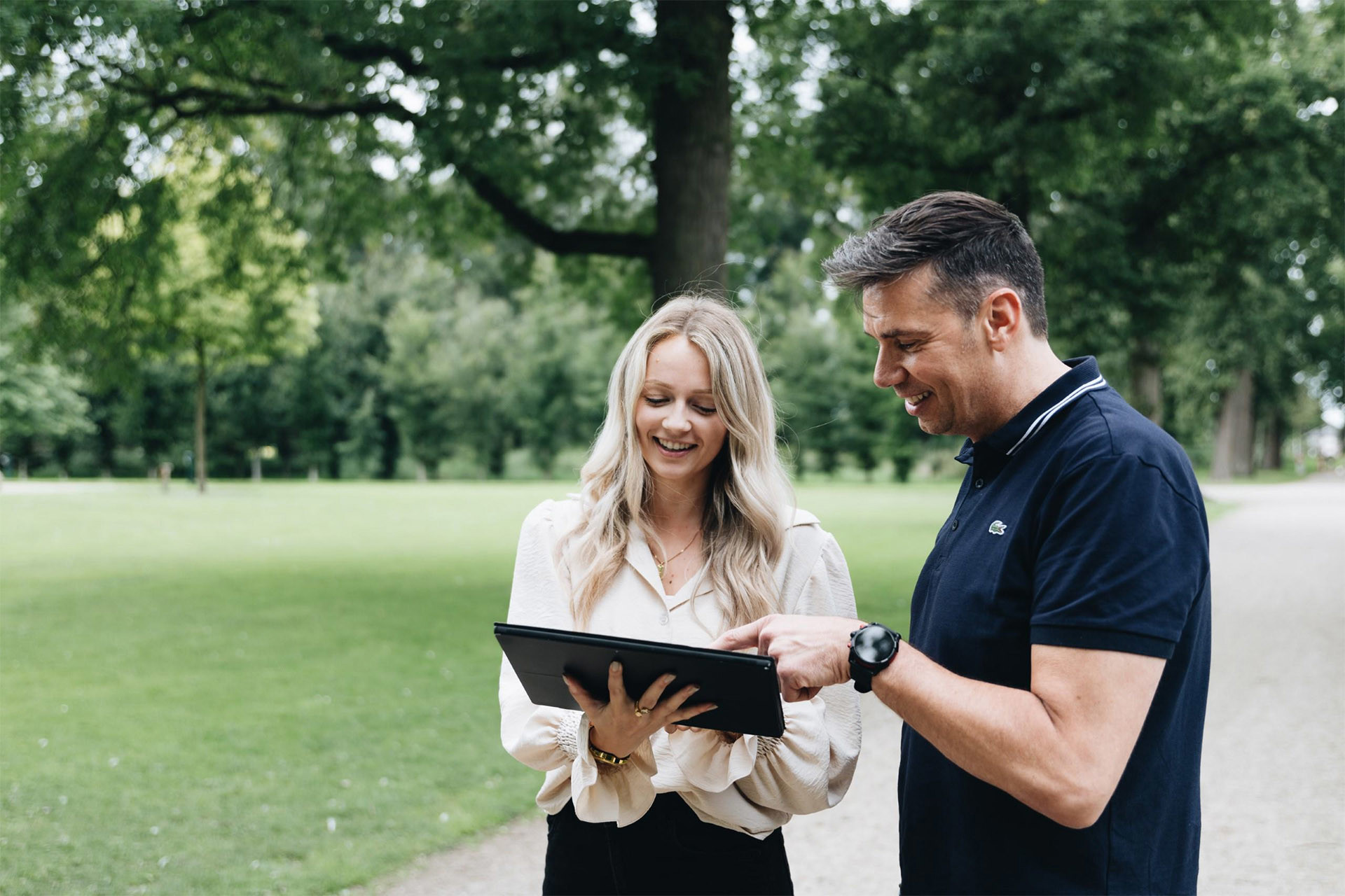Twee personen bekijken samen een tablet in een groen park, met bomen en een wandelpad op de achtergrond