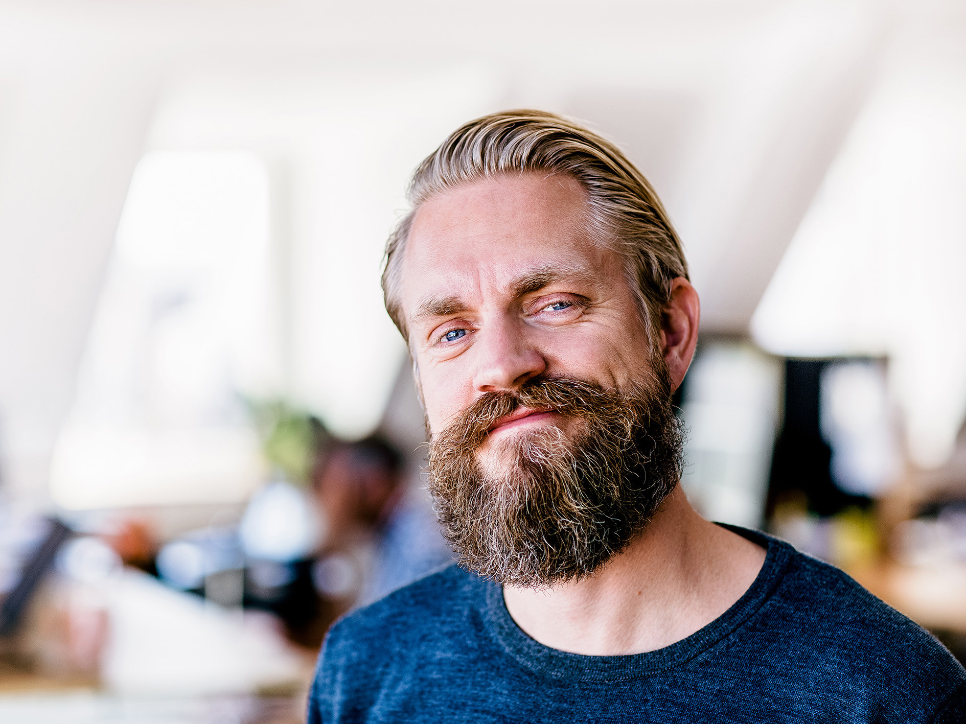 Portret van een man met baard in een kantooromgeving, met collega’s op de achtergrond