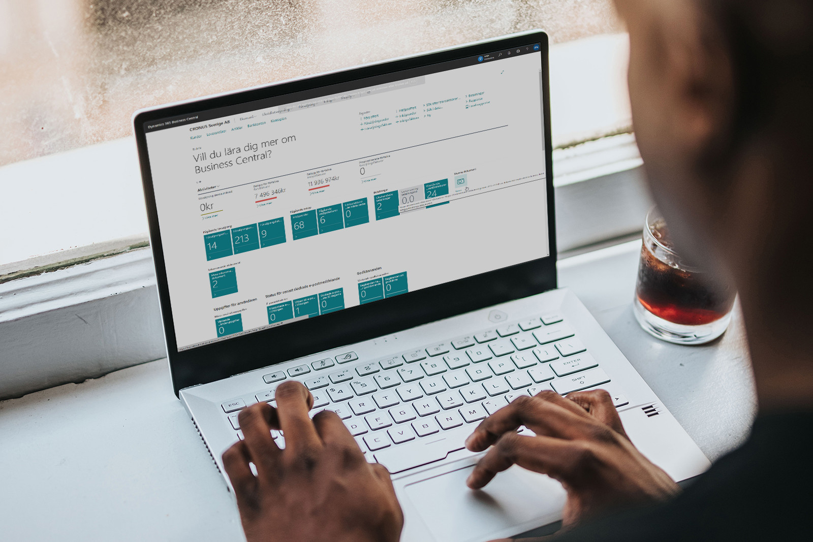 Person arbetar på en bärbar dator med Microsoft Business Central på skärmen, med ett glas dryck bredvid