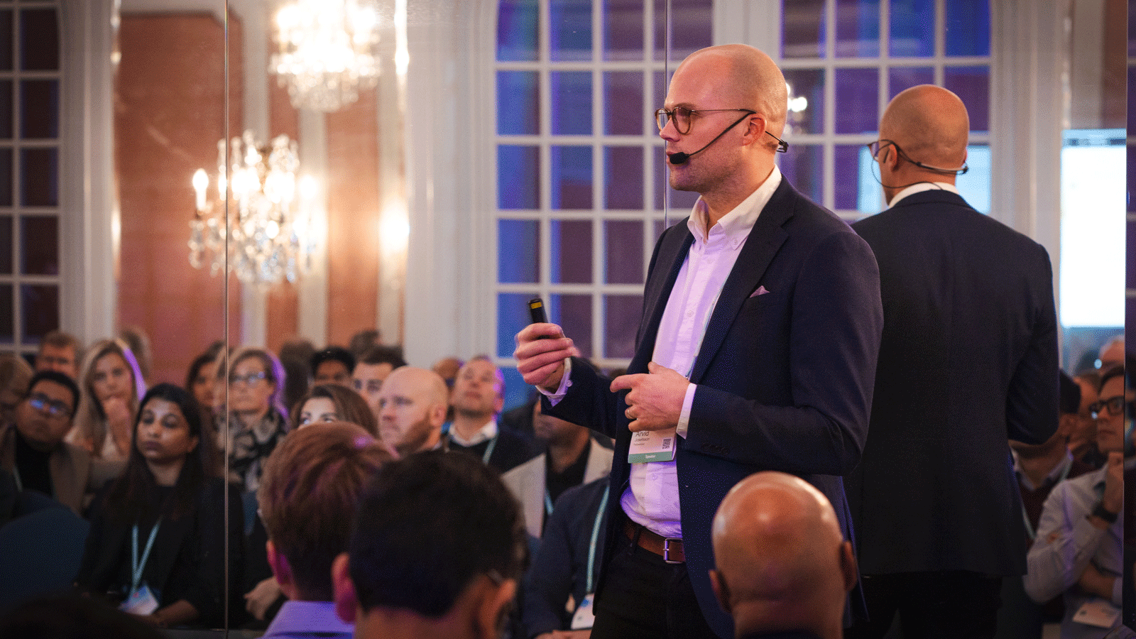 Två män i kostym talar inför publik på ett konferensmöte i en elegant sal med stora fönster och kristallkrona