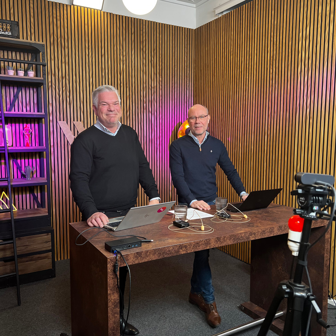 Två män står vid ett träbord med laptops i en studio med akustikpaneler och kamerautrustning