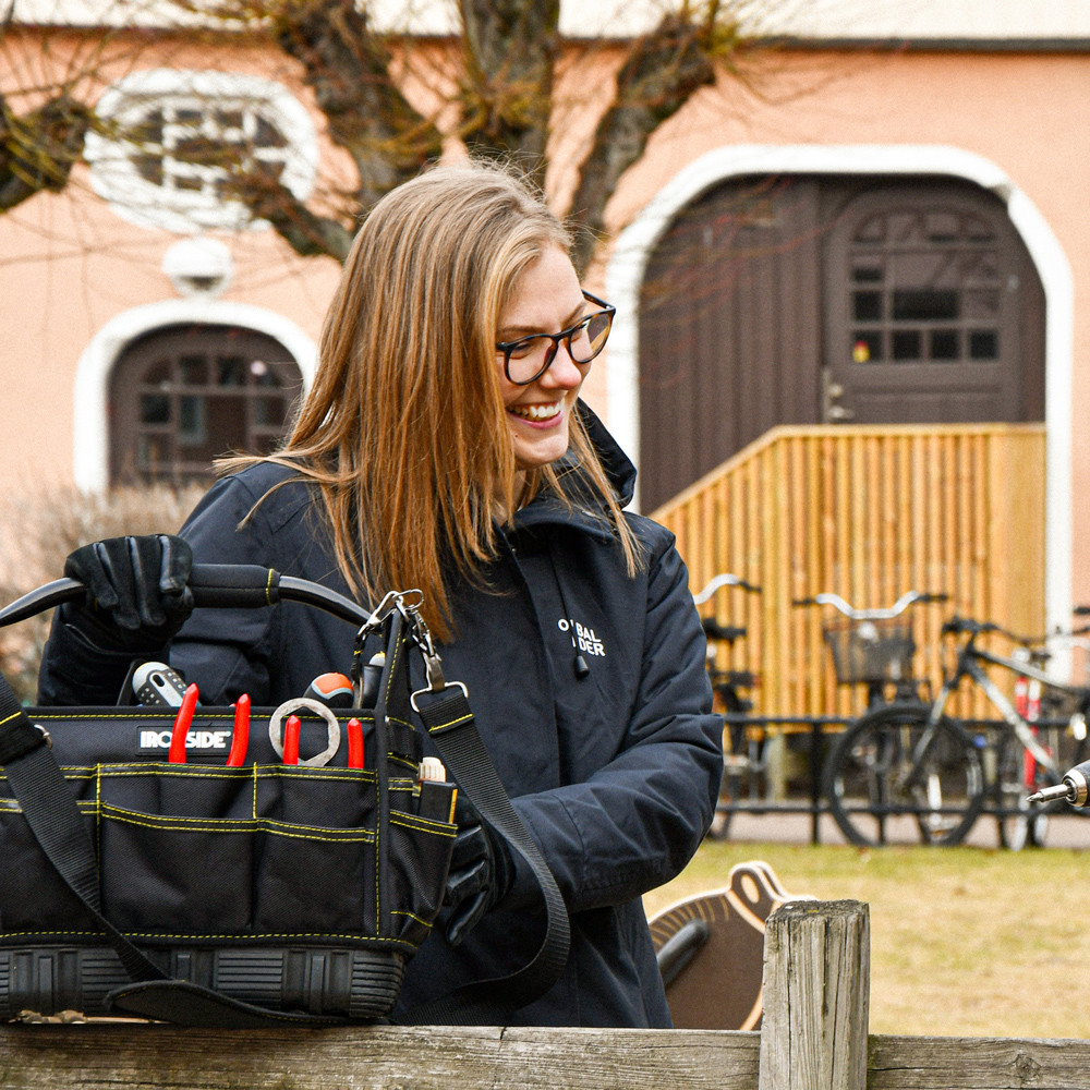 Person med verktygsväska och arbetskläder utomhus framför en byggnad, med cyklar i bakgrunden