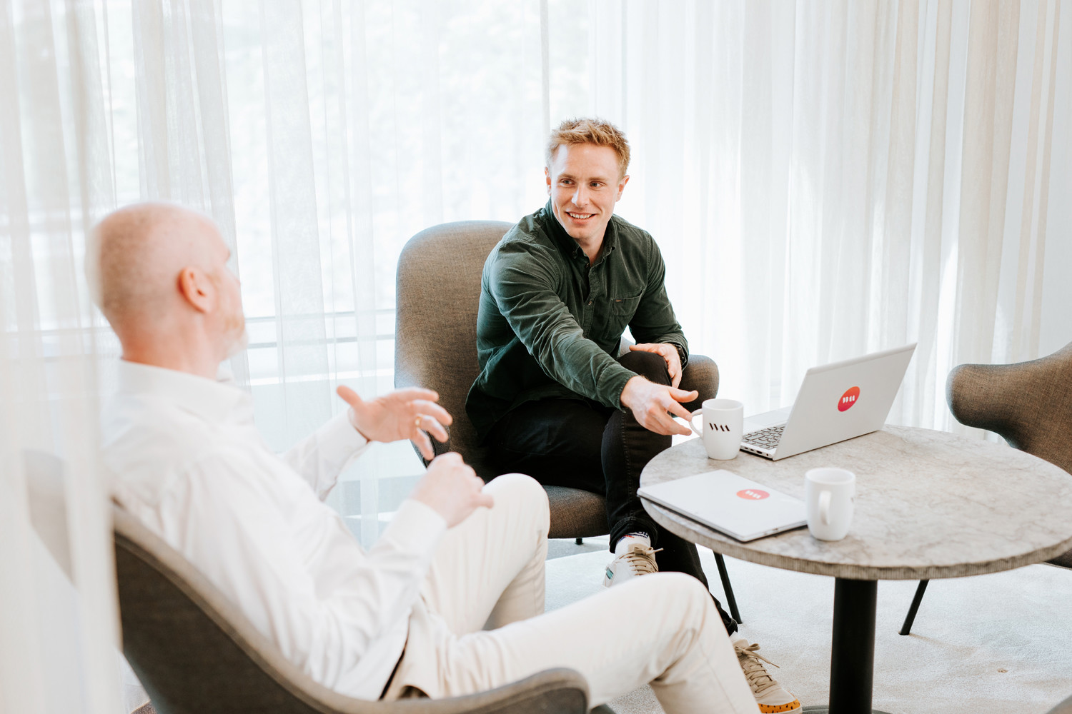 Två personer samtalar i ett ljust mötesrum med laptop och kaffekoppar, vid ett runt bord
