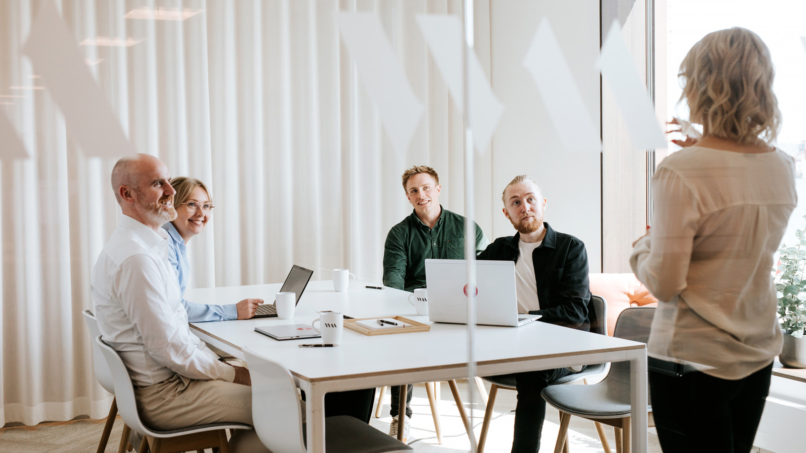 Möte i ett ljust konferensrum med flera personer runt ett bord och bärbara datorer. En kvinna står framför gruppen och presenterar