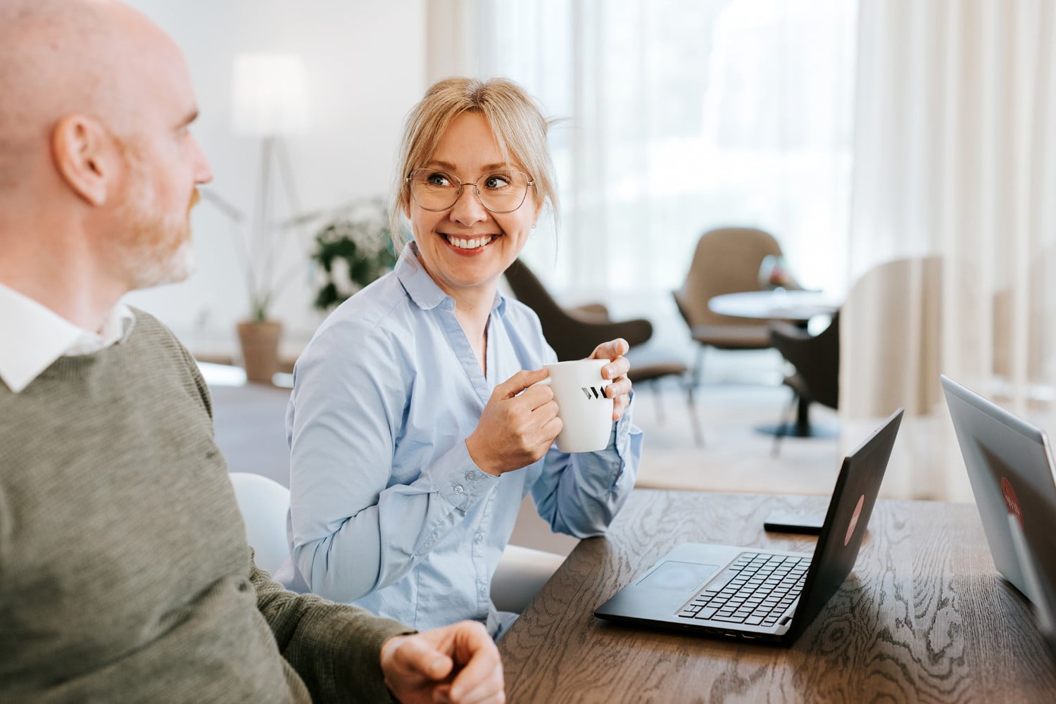 A woman is sitting in front of a computer with a cup in her hands, smiling at a male colleague